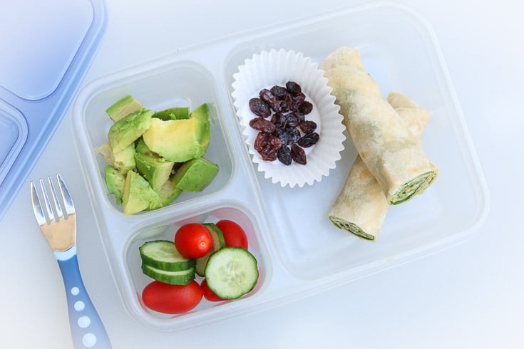 Overhead photograph of easy school lunch idea consisting of cream cheese spinach roll-ups, raisins, avocado, and raw veggies. 